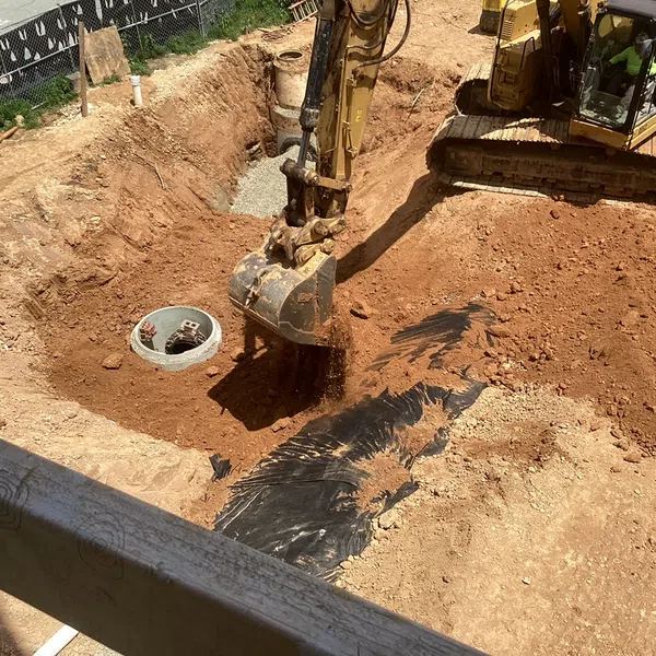 Excavator digging soil at construction site near concrete drainage pipe under sunny sky.