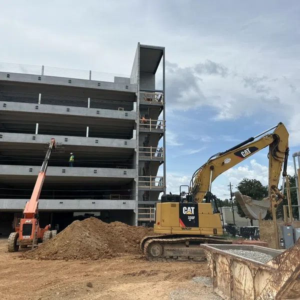 Construction site with a concrete multi-level parking structure, excavator, and crane under a partly cloudy sky.