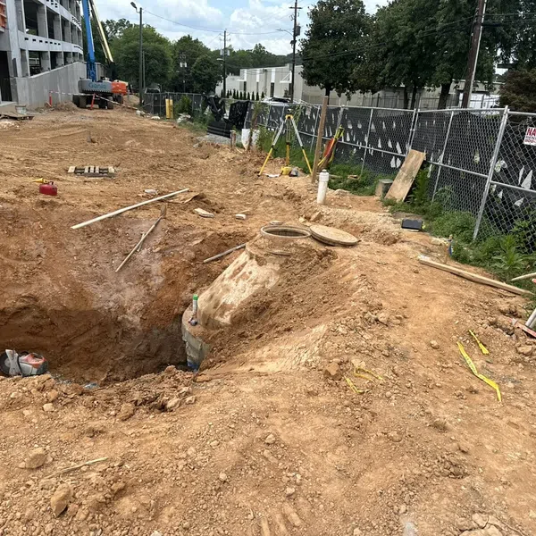 Construction site showing large excavation pit, ladders, pipes, and fenced perimeter during daytime.