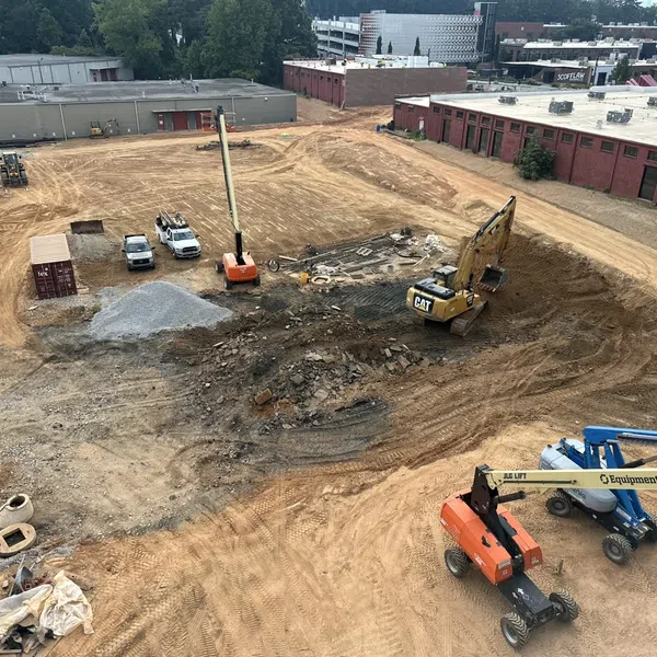 Aerial view of a construction site with excavators, dirt piles, trucks, and nearby buildings under development.