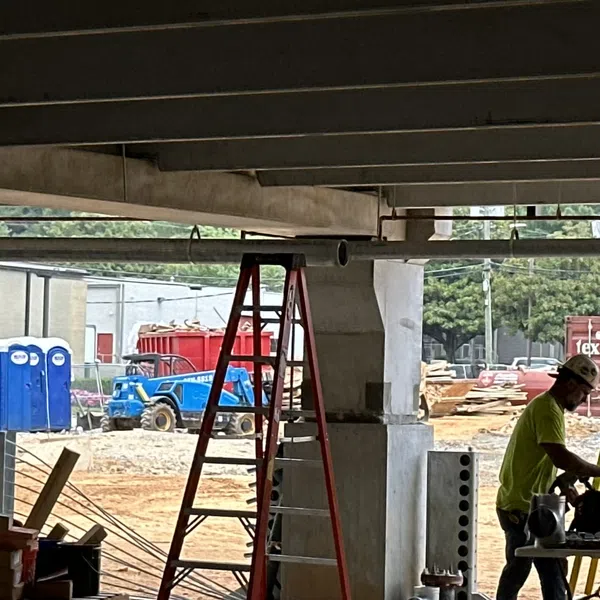 Construction site with workers in safety gear, red ladder, blue portable toilets, and equipment under a concrete structure