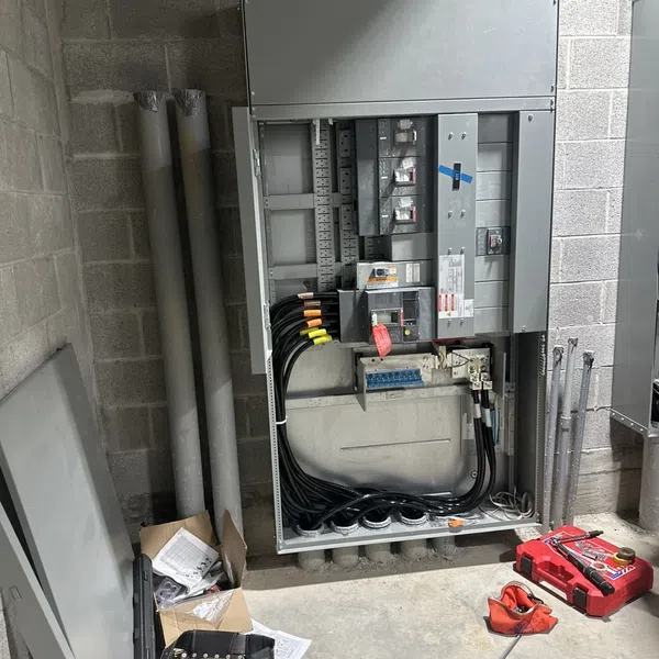 Open electrical panel with multiple circuit breakers and thick black cables in an industrial setting with tools and boots nearby