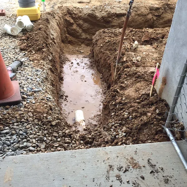 Construction site trench with exposed drainage pipe, gravel, and orange safety cone near concrete and chain link fence.