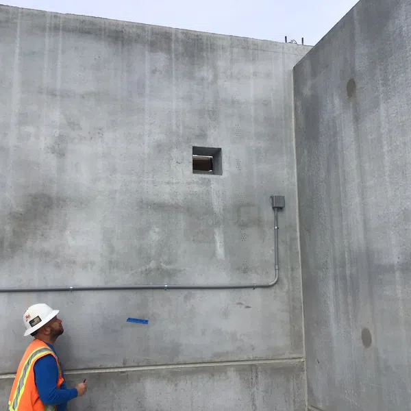 Construction worker in safety gear inspecting large gray concrete walls outdoors under cloudy sky.