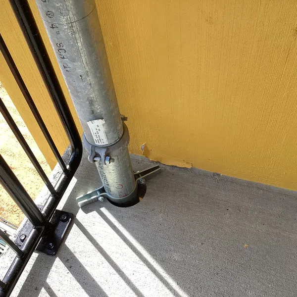 Metal construction scaffold pole clamped to a concrete balcony floor against a yellow wall and black railing.