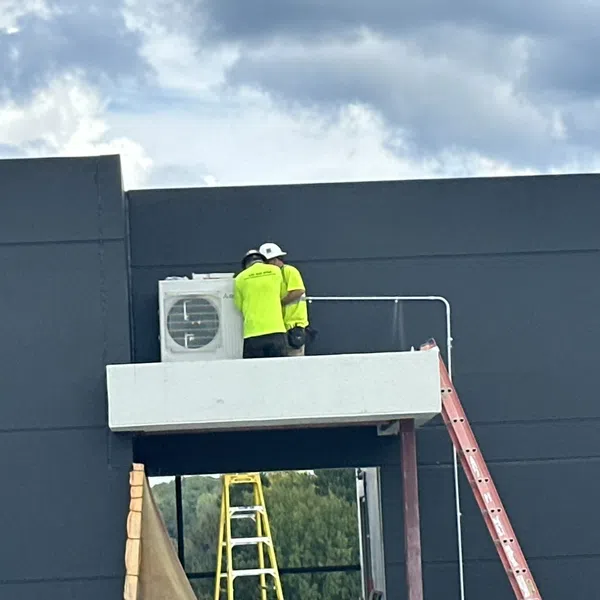 Two workers in neon shirts installing an air conditioning unit on a balcony outside a dark gray building under a cloudy sky.