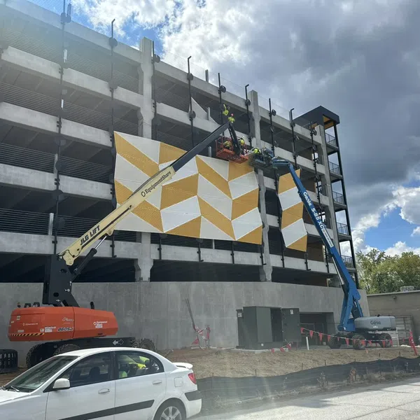 Workers install large yellow and white geometric panels on concrete parking garage using lifting equipment.