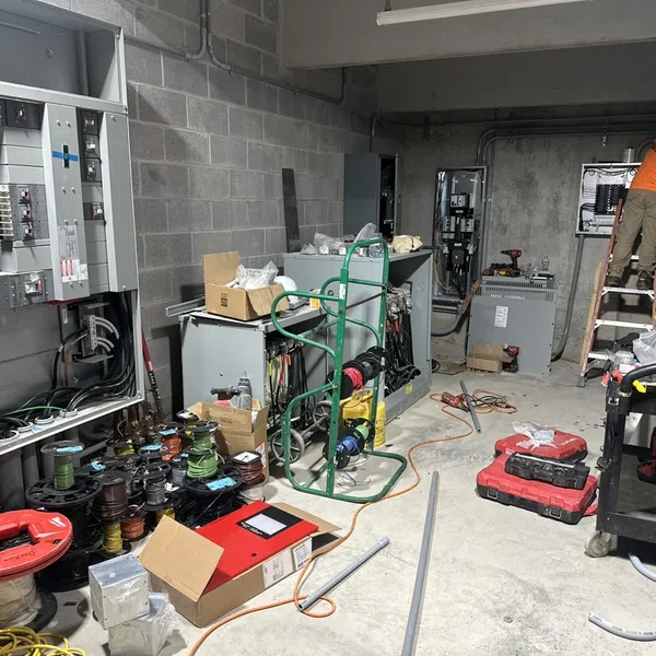 Electricians working on circuit breaker panels with wiring tools and equipment in a concrete utility room.