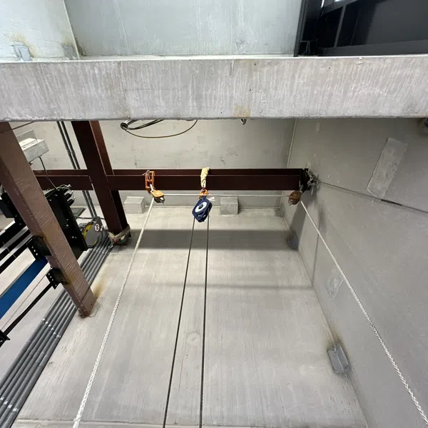 Industrial concrete shaft with metal beams, pulley system, cables, and overhead machinery in a large utility room.