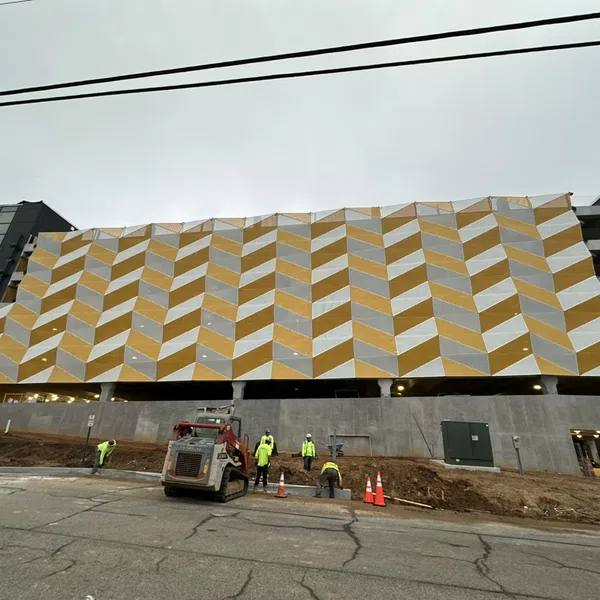 Construction workers and machinery in front of a building with a geometric yellow and white facade under cloudy sky.