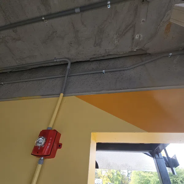 Red fire alarm with light mounted on yellow wall near an open window under a concrete ceiling with pipes.