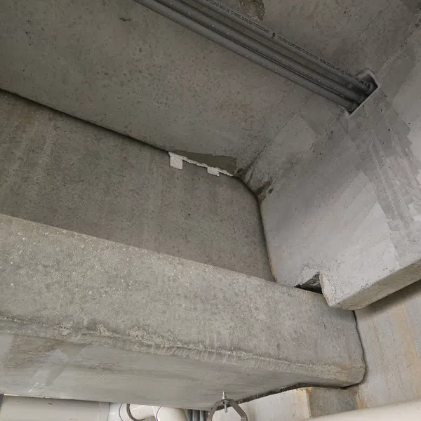 Concrete ceiling and beam with PVC pipes and metal clamps in industrial or basement setting.