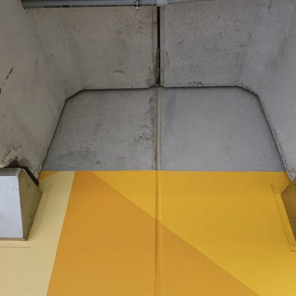 Concrete wall with pipes above and a floor painted in yellow geometric patterns in a parking garage.