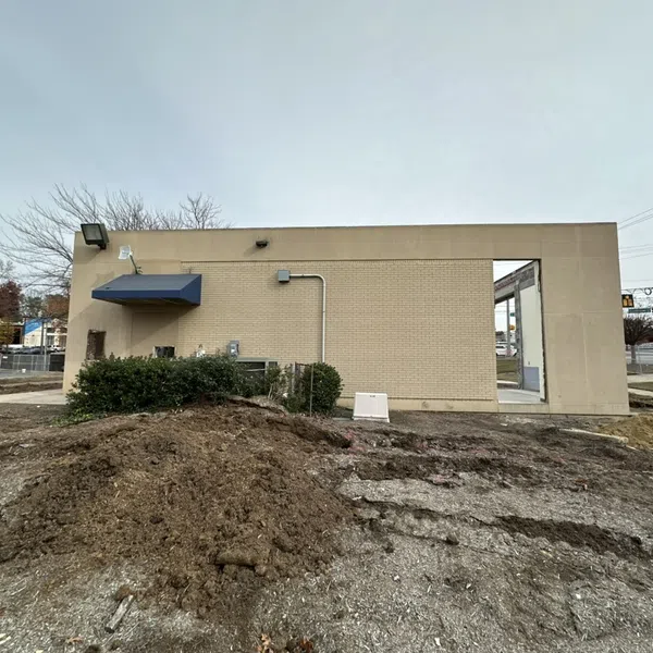 Construction site with exposed dirt mound in front of a beige brick building under cloudy sky.