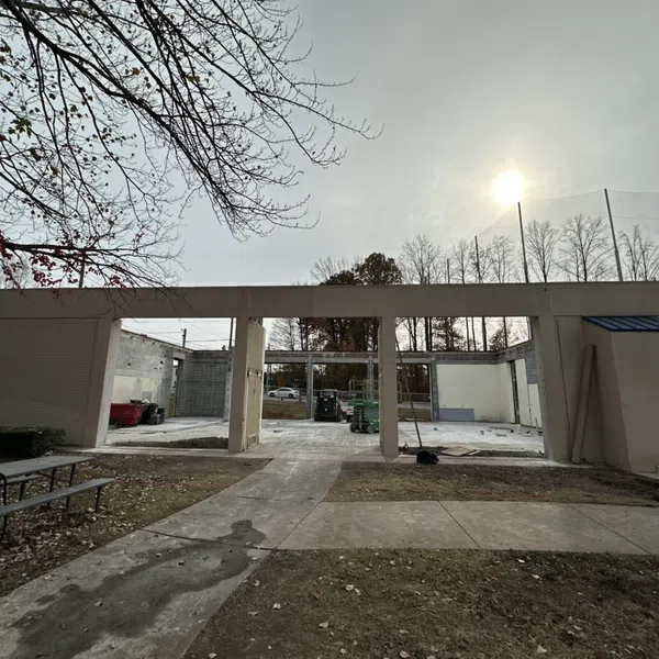 Partially constructed building with open walls under a cloudy sky, surrounded by bare trees and outdoor seating.