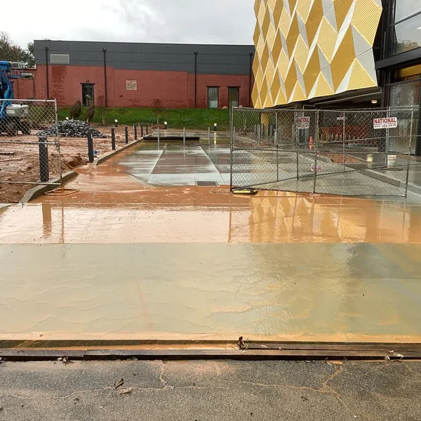 Wet concrete pavement under construction next to modern building with fencing and overcast sky