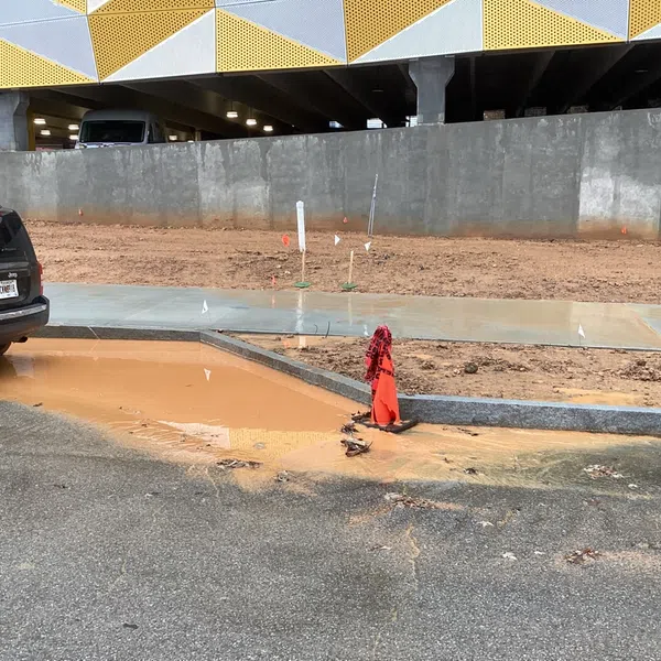 Construction site with muddy water pooling at curbside near parked black SUV and building with yellow patterned panels.