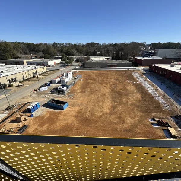 A wide view of a cleared construction site with surrounding warehouses and a perforated yellow metal railing in foreground.