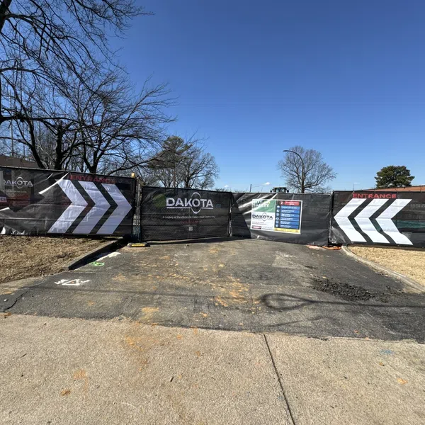 Construction site entrance blocked by fencing with Dakota signage and large white directional arrows under a clear blue sky.