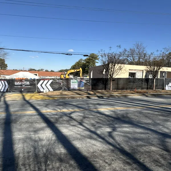Construction site with fencing and equipment behind a road with tree shadows on a sunny day