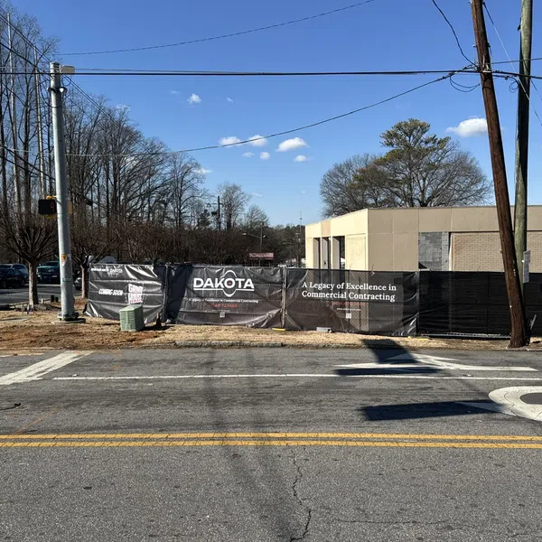 Dakota construction site with black fencing and signage on a sunny day, showing a building and leafless trees.