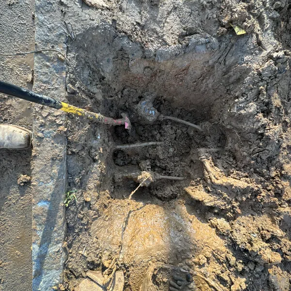 Shovel digging into muddy soil around exposed underground pipes near a concrete edge on a construction site.