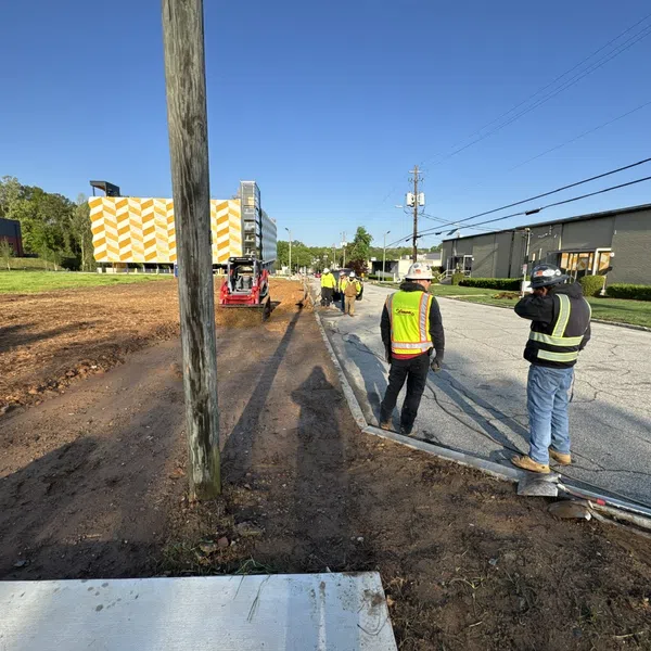 Construction workers in safety gear working on roadside excavation near utility poles under clear blue sky.