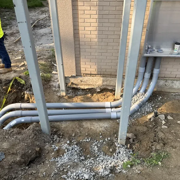 Outdoor electrical conduit installation with multiple gray pipes connected to a metal electrical box mounted on a brick wall.