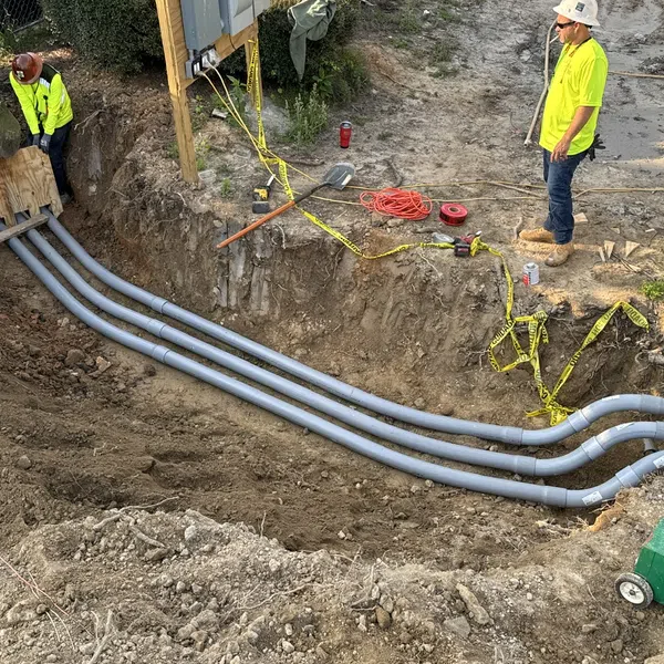 Two construction workers installing gray underground conduit pipes in a trench on a worksite.