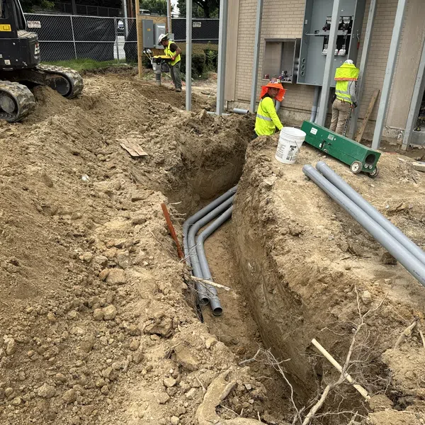 Construction workers install underground electrical conduits in a deep trench at a building site with heavy machinery.