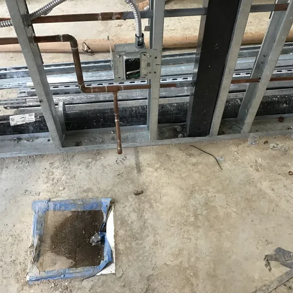 Construction site interior with exposed metal framing, pipes, and concrete floor with debris and an open floor panel.