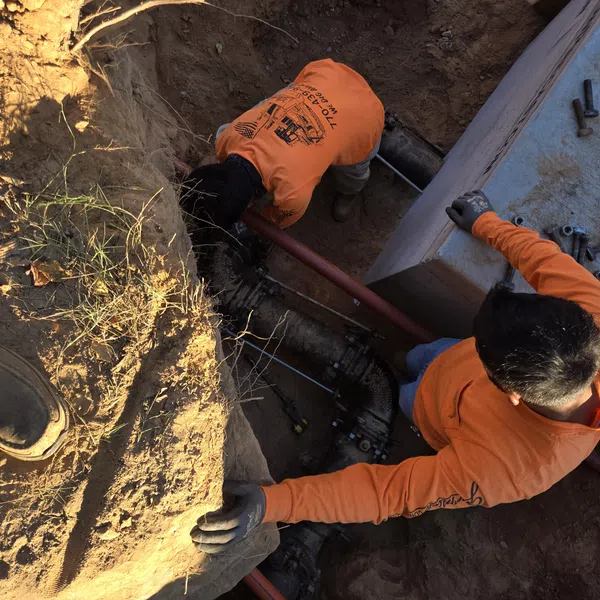 Two workers in orange shirts repairing underground pipes inside a dirt trench with tools and bolts nearby.