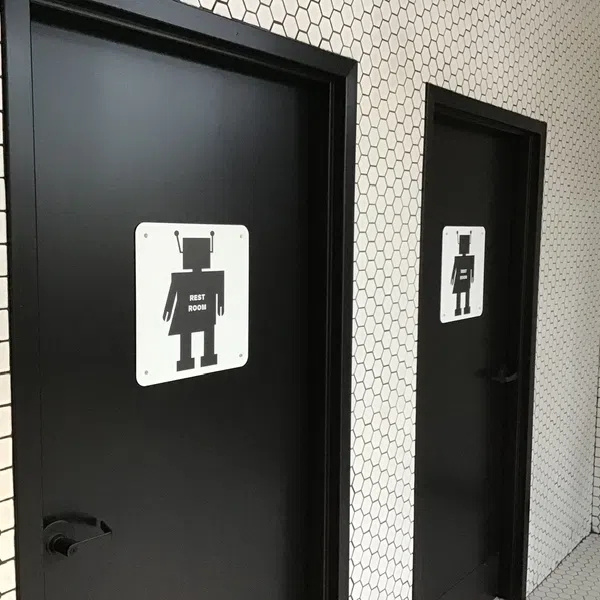 Two black restroom doors with robot icons against a white hexagonal tiled wall.