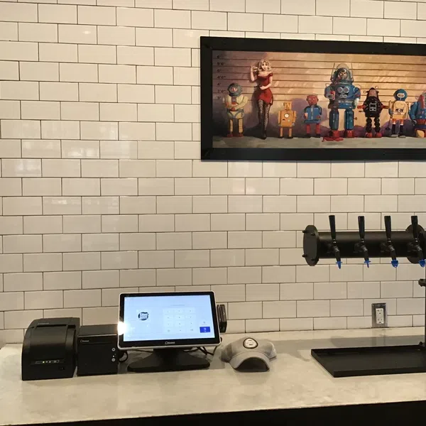Modern cafe counter with digital cashier system, beer taps, and robot-themed artwork on white tile wall.