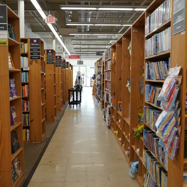 Aisle view of a bookstore with wooden shelves filled with children’s books and educational materials under bright lighting.