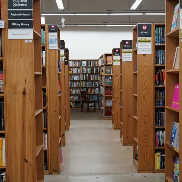 Aisle lined with wooden bookshelves in a bookstore, featuring sections for Political Science, Fiction, and Mystery books.