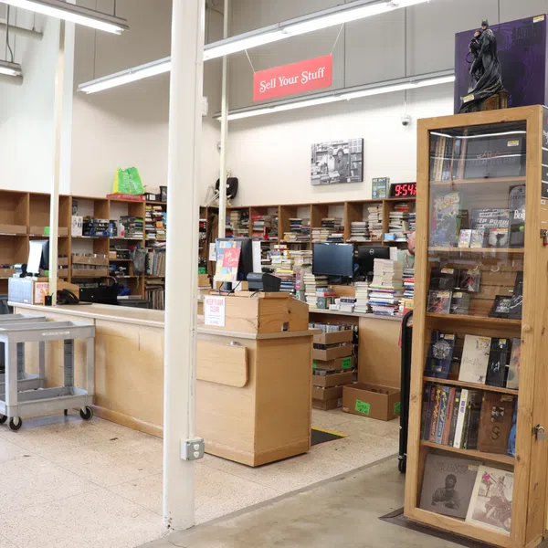 Inside a used bookstore with wooden shelves, stacks of books, checkout counters, and signs for selling and gifts section