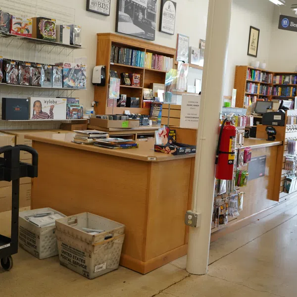 Interior of a bookstore featuring wooden counters, bookshelves, mail bins, and a black utility cart.