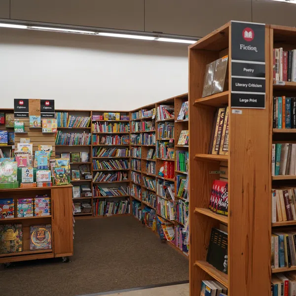 Spacious bookstore section with wooden shelves filled with fiction, poetry, children's books, and large print titles.