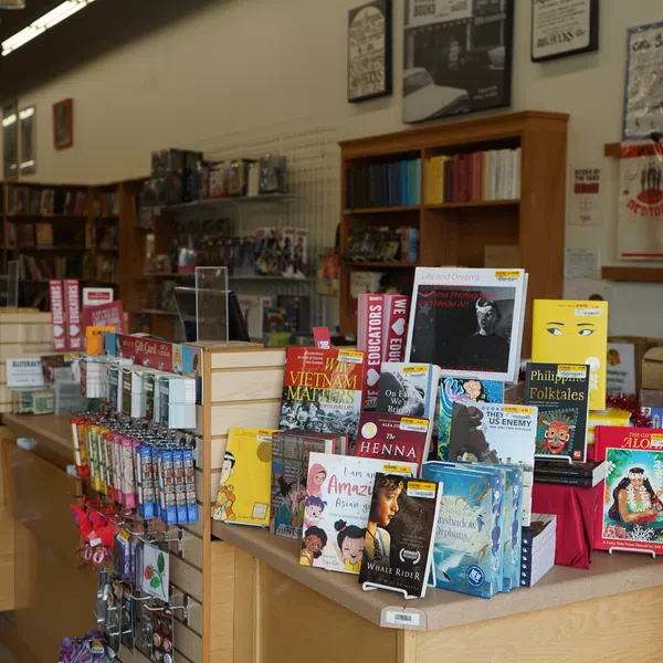 Bookstore display table showcasing diverse books, postcards, and collectibles with shelves and posters in the background.