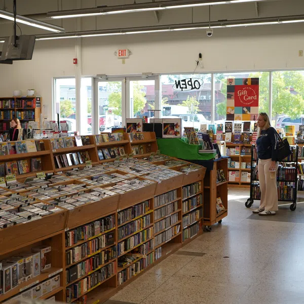 Sunny bookstore interior with wooden shelves full of books and a woman browsing near the entrance with glass doors.