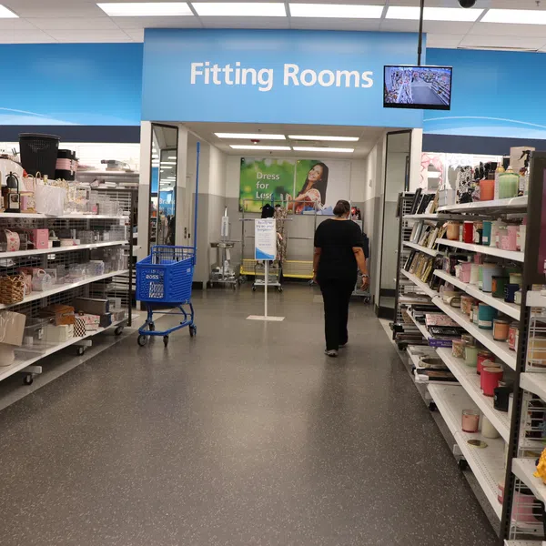 Inside a retail store aisle leading to fitting rooms with shelves of home goods on both sides and a shopper walking ahead.