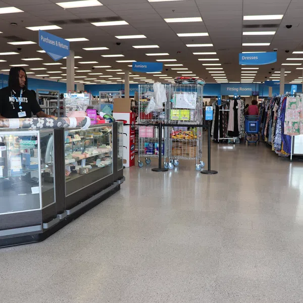 Inside a spacious thrift store with sections for juniors, dresses, and fitting rooms, and a staff member at a glass counter.