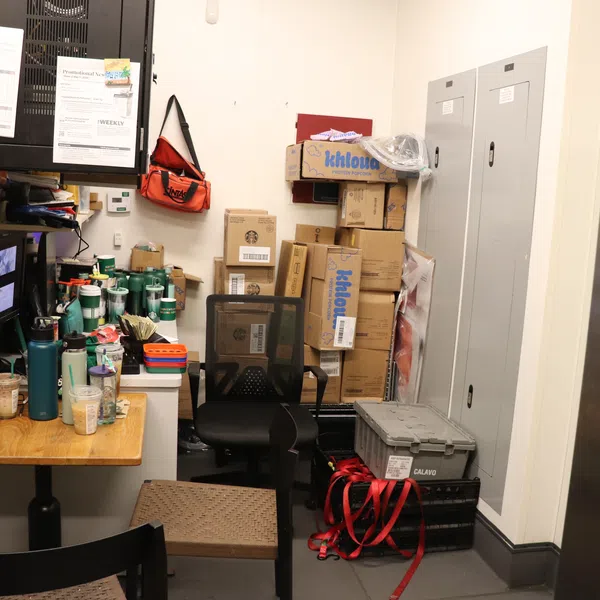 Cluttered small room with stacked boxes, office chair, table with cups and containers, security monitors on wall.