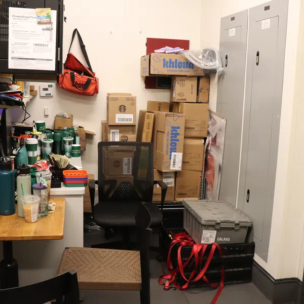 Small cluttered storage room with stacked boxes, office chair, table with cups, storage bins, and security monitors.