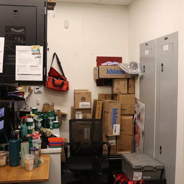 Small office or storage room cluttered with boxes, reusable cups, chairs, and office supplies against white walls.