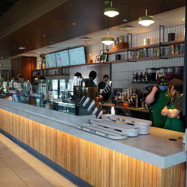 Modern coffee shop interior with wooden counter, overhead lights, menu screens, and baristas in green aprons.
