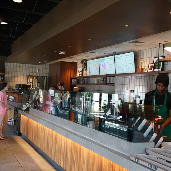 Modern coffee shop interior with wooden counter, digital menu screens, baristas preparing drinks, and a customer in pink outfit