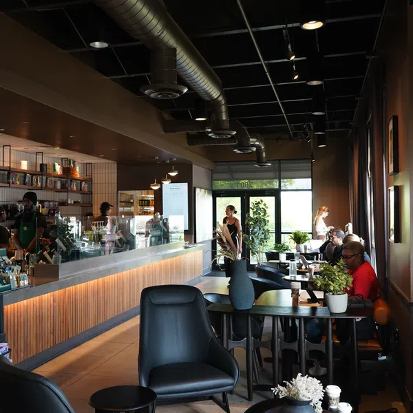 Modern coffee shop interior with customers, wooden counter, black chairs, and industrial ceiling ductwork.