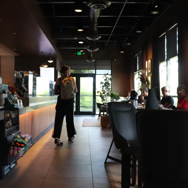 Person with backpack walking inside a modern coffee shop with customers seated by the window and ambient lighting.